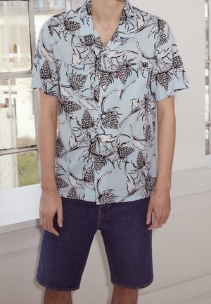 Homme portant une chemise bleu clair à manches courtes avec un motif floral noir et blanc et un short en jean bleu foncé, debout à l'intérieur.