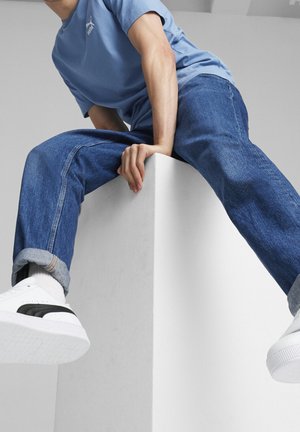 Personne portant un T-shirt Puma bleu, un jean bleu et des baskets blanches, assise sur un cube blanc, tenant le bord d'une main.