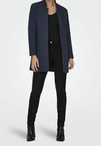 Cappotto testurizzato blu navy con taglio dritto, senza colletto e maniche lunghe, indossato sopra un top nero e pantaloni neri aderenti. Stivaletti neri.