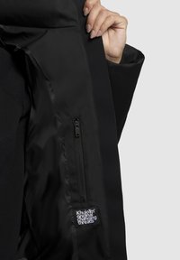 Hand hält eine offene schwarze Jacke, die eine innere Zipptasche und ein weißes Etikett mit dem Text "Khujo original changing threads" zeigt.