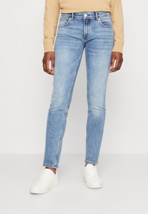 Persona con vaqueros ajustados azul claro, camiseta de manga larga beige y zapatillas blancas, de pie sobre un fondo blanco liso.