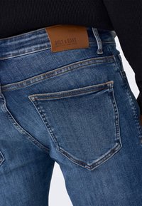 Vaqueros de mezclilla azul oscuro con un diseño clásico de cinco bolsillos, con una parche de cuero en la parte trasera de la cintura y detalles de costura visibles.