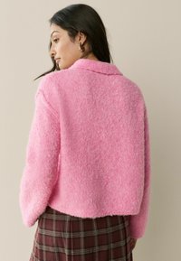 Rožinė pūkuota striukė su koloriu ir per dideliu siluetu, derinama su ruduošiu raštuota sijonu. Tekstūruotas audinys suteikia švelnų vaizdą.