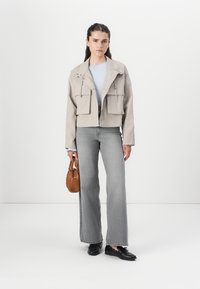 Veste beige cropped avec deux poches avant, portée sur un top à côtes bleu clair. Un pantalon en jean gris à jambes larges et des chaussures noires complètent la tenue.