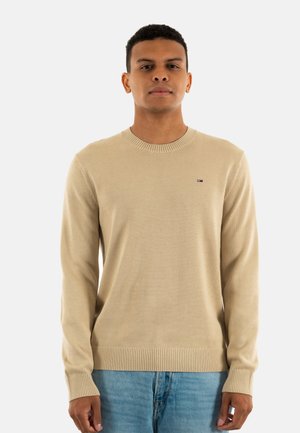 Jersey de punto color beige con cuello redondo, puños y dobladillo acanalados. Presenta un pequeño logo en el pecho. El modelo lleva unos jeans de color azul claro.