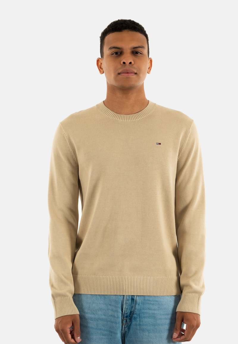 Maglione beige lavorato a maglia con collo rotondo, polsini e orlo a costine. Presenta un piccolo logo sul petto. Il modello indossa jeans azzurri chiari.