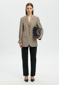 Mujer vestida con un blazer beige abotonado, pantalones negros de pierna recta, tacones negros de punta, sosteniendo un gran bolso de cuero negro bajo su brazo.
