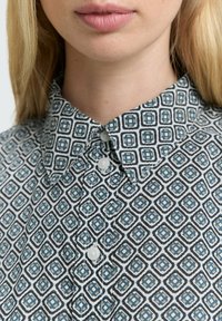 Detaily na osobě, která má na sobě vzorovanou košili s modro-černými geometrickými tvary a třemi viditelnými zapnutými knoflíky.