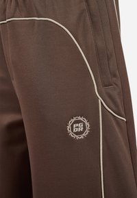 Pantalon de sport marron avec une texture lisse, comportant des passepoils beiges le long des coutures et un logo brodé avec un design circulaire sur la cuisse.