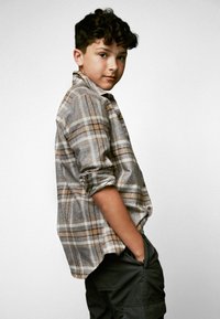 Grå och beige rutmönstrad skjorta med knappar och uppvikta ärmar, kombinerad med mörka cargobyxor. Skjortan har en avslappnad passform och mjuk textur.