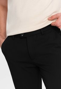 Mustat räätälöidyt housut, jotka on valmistettu sileästä kankaasta. Niissä on litteä etuosa, sivutaskut ja nappikiinnitys vyötäröllä.