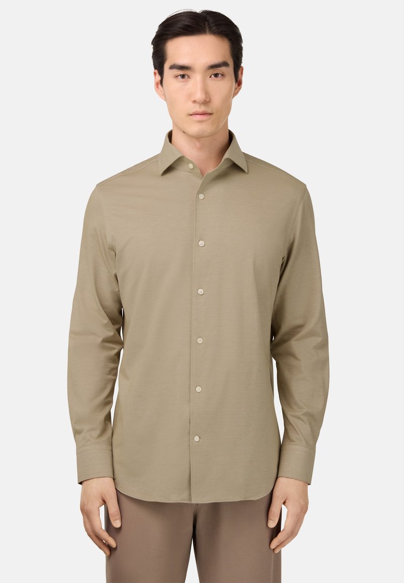 Camisa de manga larga beige con botón frontal, cuello puntiagudo, textura suave y diseño ajustado, combinada con pantalones marrones claros.