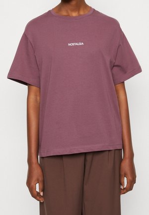 Person iført en løs mauve T-shirt med teksten "NOSTALGIA" på brystet og matchende brune bukser, stående mod en ensfarvet baggrund.