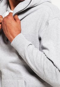 Uomo che indossa una felpa grigia chiara con zip, che tira su la zip con la mano destra.