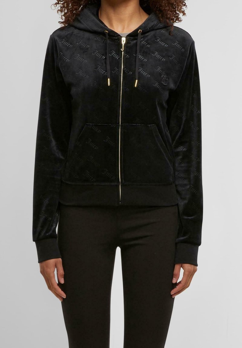 Femme portant un sweat à capuche zippé en velours noir avec des cordons et un pantalon noir assorti, debout devant un fond uni gris clair.