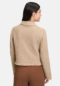 Giacca beige corta con tessuto testurizzato, ampio colletto e orlo sfrangiato. La vista dal retro mostra maniche lisce e una vestibilità casual abbinata a pantaloni marroni.