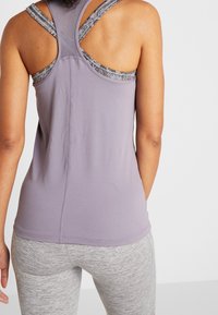 Fioletowy sportowy top bez rękawów z teksturowanej tkaniny, z plecami w stylu racerback i kontrastującymi szarymi detalami na ramiączkach. Noszony z szarymi legginsami.