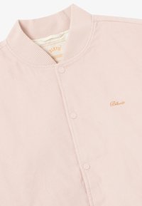 Pink corduroy jakke med rund krave, frontknaplukning og subtil logo-brodering i guld. Teksturret stof og minimalistisk design.