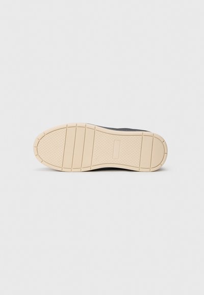 Schuhsohle mit beigem Gummi-Profil, das horizontale Rillen und ein Rautenmuster auf einem schlichten grauen Hintergrund aufweist.