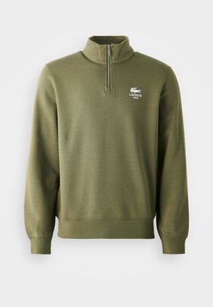 Olive zelen pulover s četrtinskim zadrgo, izdelan iz mehkega materiala, z rebrastimi manšeti in pasom, z belim logotipom Lacoste na prsih.