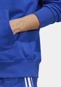 Felpa blu in morbido tessuto con tasca a marsupio, polsini e orlo a coste. Presenta una superficie liscia e un design sportivo.