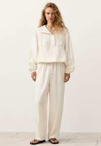 Femme debout portant un pull blanc ample avec cordons, pantalon blanc assorti et tongs noires sur un fond uni.