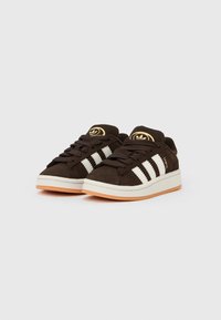 Καφέ σουέντ αθλητικά παπούτσια με λευκές ρίγες, στρογγυλή μύτη, λαστιχένια σόλα και χρυσά λογότυπα στη γλώσσα. Οι υφασμένες κορδόνες ολοκληρώνουν το σχέδιο.