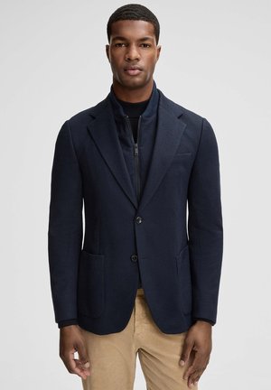 Marineblauwe blazer met twee voorzakken, enkele knoopsluiting en een gestructureerde afwerking. Voorzien van een ritssluiting aan de binnenlaag en een getailleerde pasvorm.