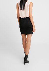 Kvinna i en ljusrosa ärmlös topp, svart strukturerad minikjol och svarta högklackade ankelstövlar, som står framför en enfärgad bakgrund.