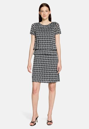 Frau steht, trägt ein schwarz-weißes kurzärmeliges gemustertes Kleid und schwarze Sandalen mit Absatz vor einem einfarbigen hellen Hintergrund.