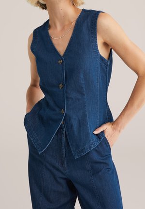 Vrouw draagt een mouwloos donkerblauw denimvest met knopen en bijpassende high-waisted denimbroek, handen in de zakken, tegen een neutrale achtergrond.
