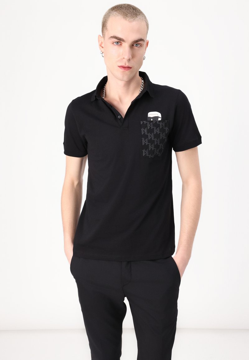 Svart polo skjorte laget av lett stoff, med et mønstret lomme, en krage og minimalistisk hvit logo-detalj.
