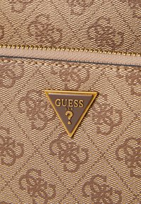 Portafoglio in tessuto marrone con texture, caratterizzato da una placca logo triangolare in tono dorato con "GUESS USA" e un motivo di loghi intrecciati.