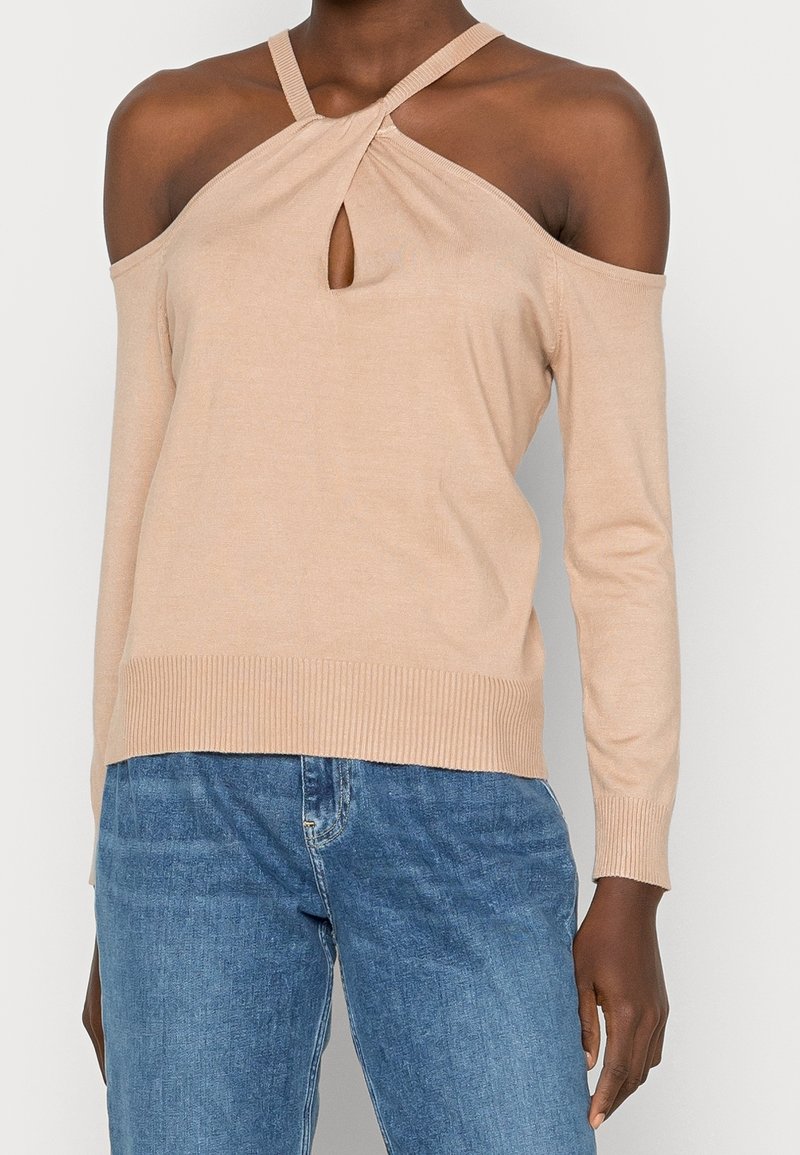 Femme portant un pull beige épaules dénudées avec un col halter torsadé et un jean bleu, debout devant un fond uni.