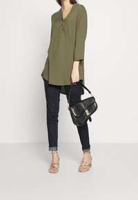 Olivgrön tunika med V-ringning och tre knappar, tillsammans med mörka jeans och sandaler med remmar i beige. Modellen håller en svart handväska.