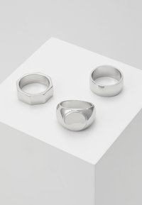 Três anéis de prata exibidos em um pedestal branco: um aro hexagonal, um design oval liso e um aro arredondado com acabamento polido.