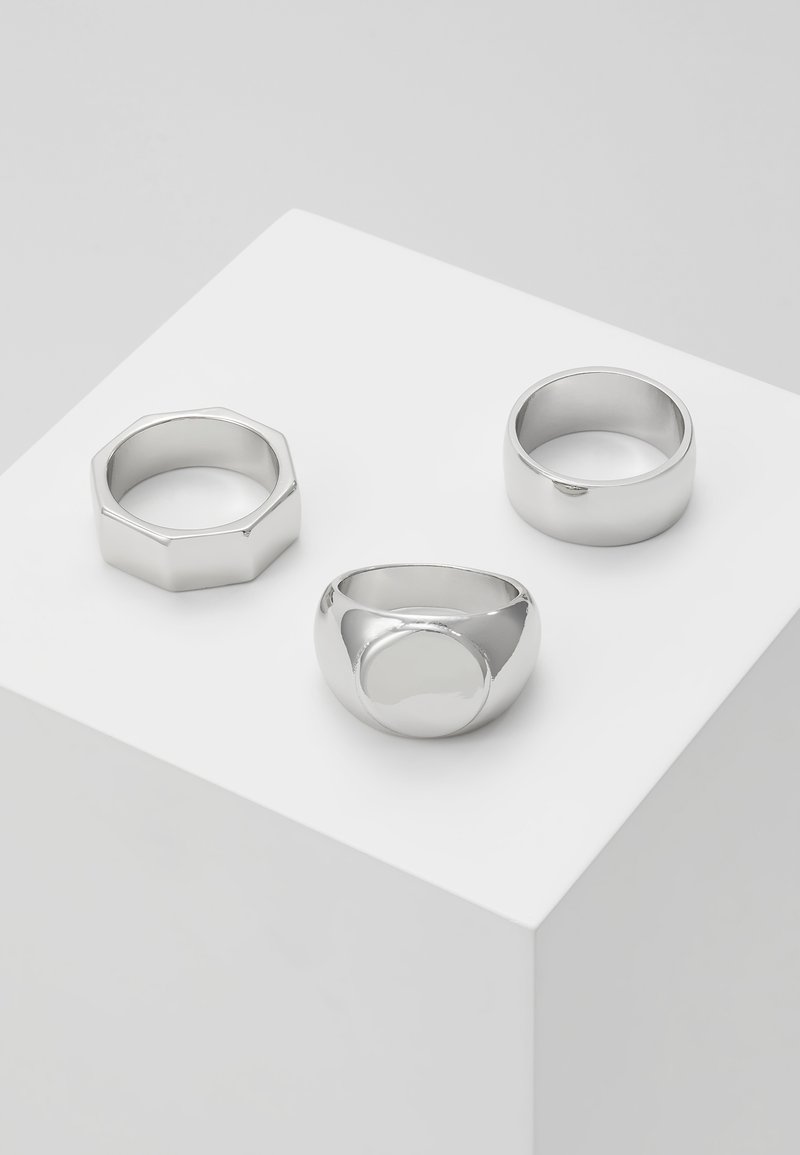 Três anéis de prata exibidos em um pedestal branco: um aro hexagonal, um design oval liso e um aro arredondado com acabamento polido.