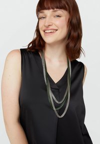 Collana a più filamenti con catene metallizzate verdi e argento, abbinata a una maglia nera senza maniche e con scollatura a V.