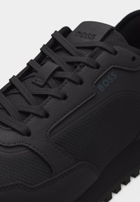 Baskets noires texturées avec des lacets noirs comportant le nom de la marque Boss sur la languette et le côté près des lacets.