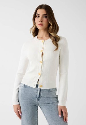 Frau mit langen, welligen Haaren, die einen weißen, geknöpften Cardigan mit goldenen Knöpfen und hellblaue Jeans trägt, steht vor einem einfarbigen Hintergrund.