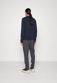 Marinblå tröja, lättstickad; grå rutiga byxor med avslappnad passform; bruna och krämfärgade sneakers; modell med långt hår, sedd bakifrån.