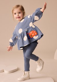 Niña joven equilibrándose sobre un pie, vistiendo un vestido azul con lunares y un parche de mariquita naranja, leggings azules y zapatillas blancas de caña alta.