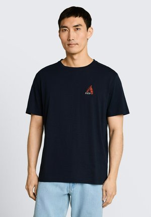 Mężczyzna noszący jednolitą czarną koszulkę z małą czerwoną grafiką na lewej piersi, w zestawieniu z jasnoniebieskimi dżinsami na jednolitym tle.