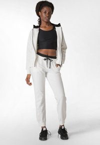 Witte zip-up jas met zwarte accenten, gecombineerd met een zwart crop top en witte joggerbroeken met een trekkoord taille en zwarte afwerking. Zwarte sneakers.