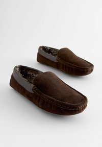 Chaussons en daim marron avec un accent en cuir lisse sur les côtés, présentant une doublure intérieure douce et pelucheuse ainsi que des coutures décoratives le long des bords.