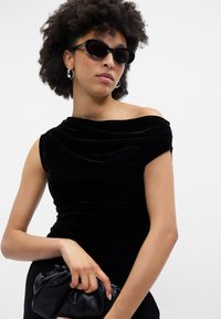 Μαύρο βελούδινο φόρεμα off-shoulder με draped λεπτομέρεια. Το μοντέλο κρατάει μία μικρή μαύρη τσάντα, φοράει μαύρα οβάλ γυαλιά ηλίου και ασημένια σκουλαρίκια κρίκους.