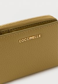 Vihertävän ruskea vetoketjullinen nahkalompakko, jossa on teksturoitu pinta ja kultainen "Coccinelle"-logo. Sileä vetoketju kultasävyisillä metalliosilla.