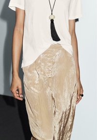 Beige, glanzende, brede broek met een gestructureerd oppervlak, gecombineerd met een losvallend wit T-shirt en een lange, zwarte kwastketting.