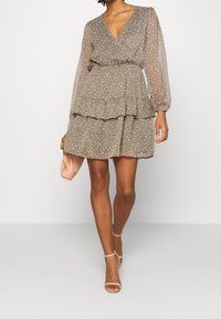 Robe en chiffon olive à motif floral, avec un décolleté en V, de longues manches bouffantes, des volants superposés, une taille cintrée, portée avec une pochette rose et des sandales.