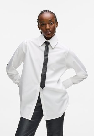 Balta garām piedurknēm krekls ar norādītu apkakli un melnu kaklasaiti, ar pogām un vaļīgu piegriezumu, valkāts ar melniem pieguļošiem biksēm.